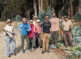 A group of people with garden tools, standing in a cultivated area with trees in the background.