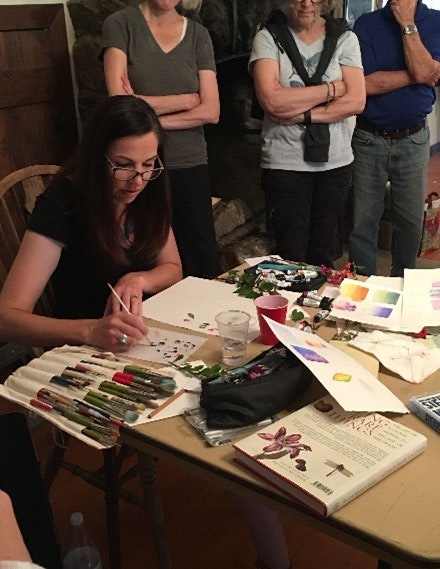 A woman painting at a table with onlookers. Art supplies scattered around.