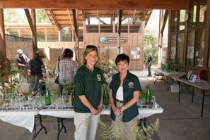 Two people in uniforms standing by a table with plants, indoors at an event or exhibition.