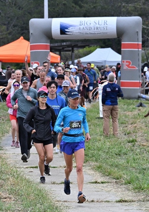 A group of runners is participating in a race near a Big Sur Land Trust banner, with many spectators and tents in the background.