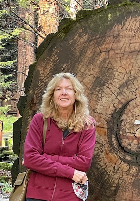 A smiling woman in a maroon jacket standing in front of a large tree cross-section.