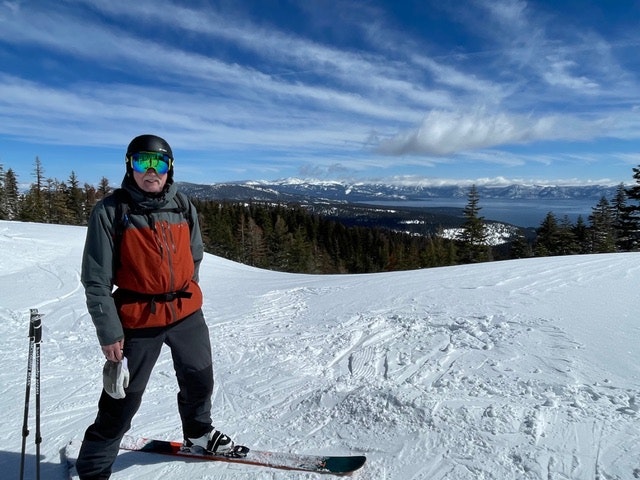 Person in ski gear on snowy slopes with ski poles and trees under a clear blue sky.