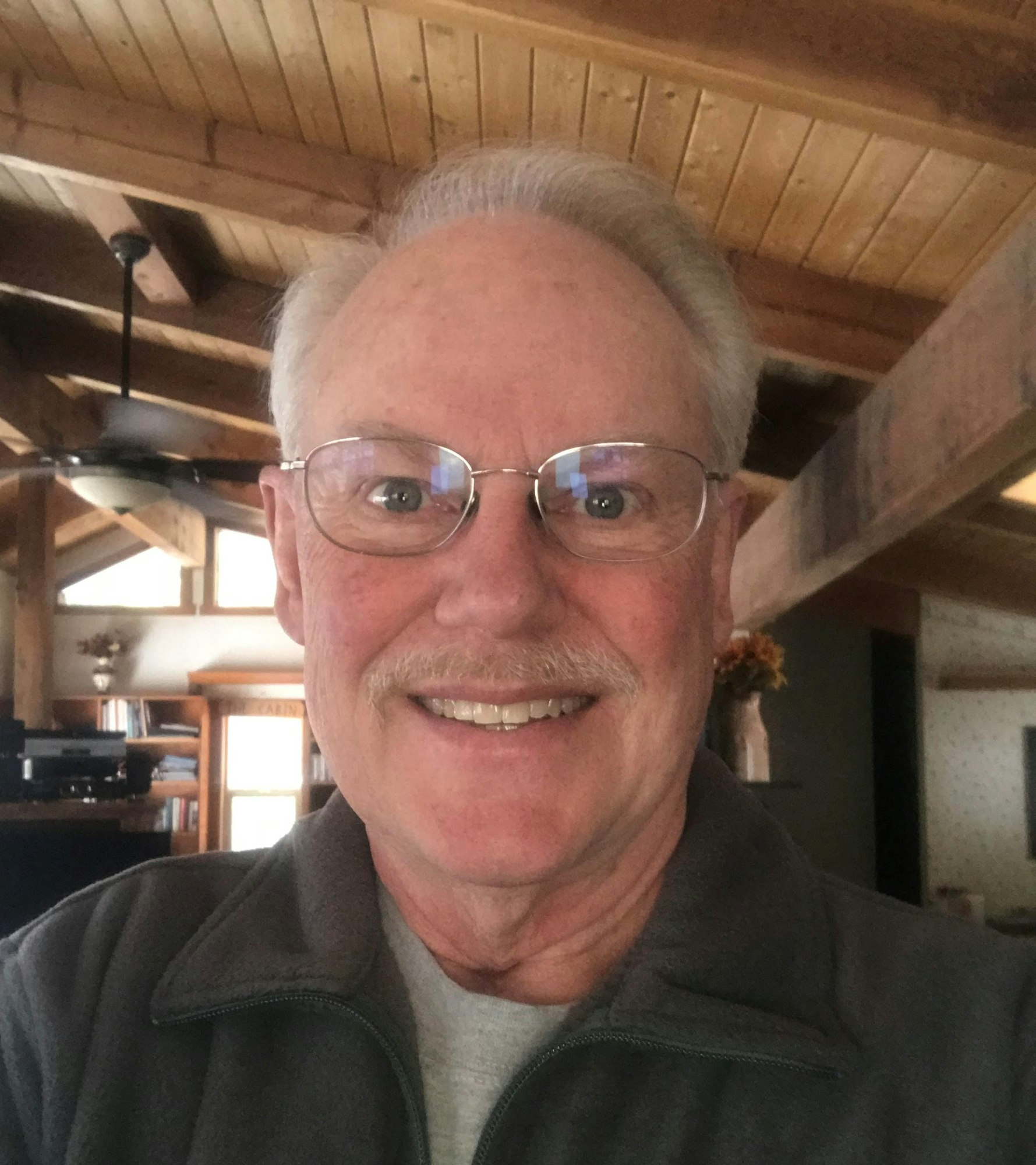 A smiling man with glasses and a mustache poses for a selfie in a cozy, wooden interior.