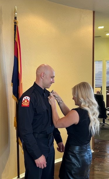 A woman pins a badge on a man in uniform beside a flag indoors.