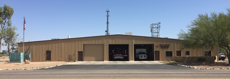 A large, tan metal building with two garage doors, housing fire trucks, an American flag, and communication towers nearby.