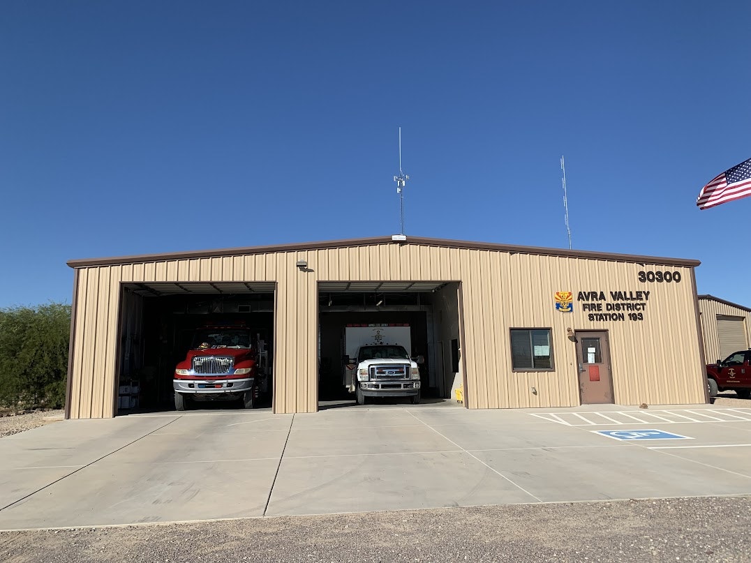 A fire station building with two fire trucks visible, featuring the Avra Valley Fire District sign and an American flag.