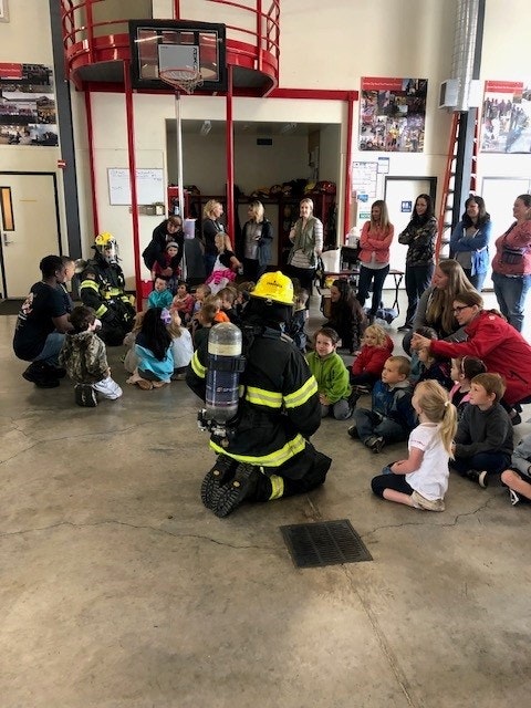 A firefighter is demonstrating equipment to an engaged group of children and adults inside a fire station.