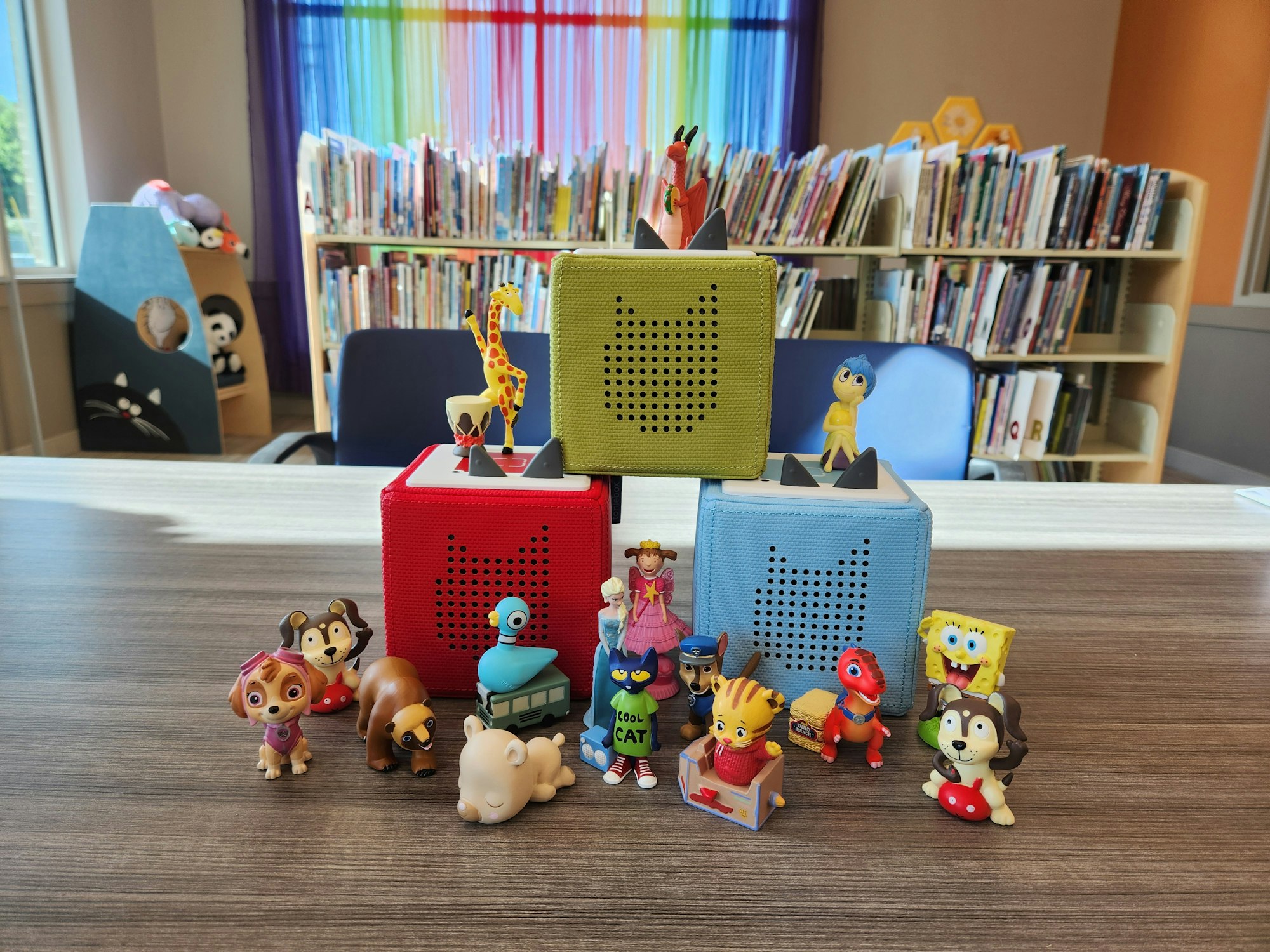 Toy figures and colored audio boxes on a table in a library with bookshelves and cat-themed decor in the background.