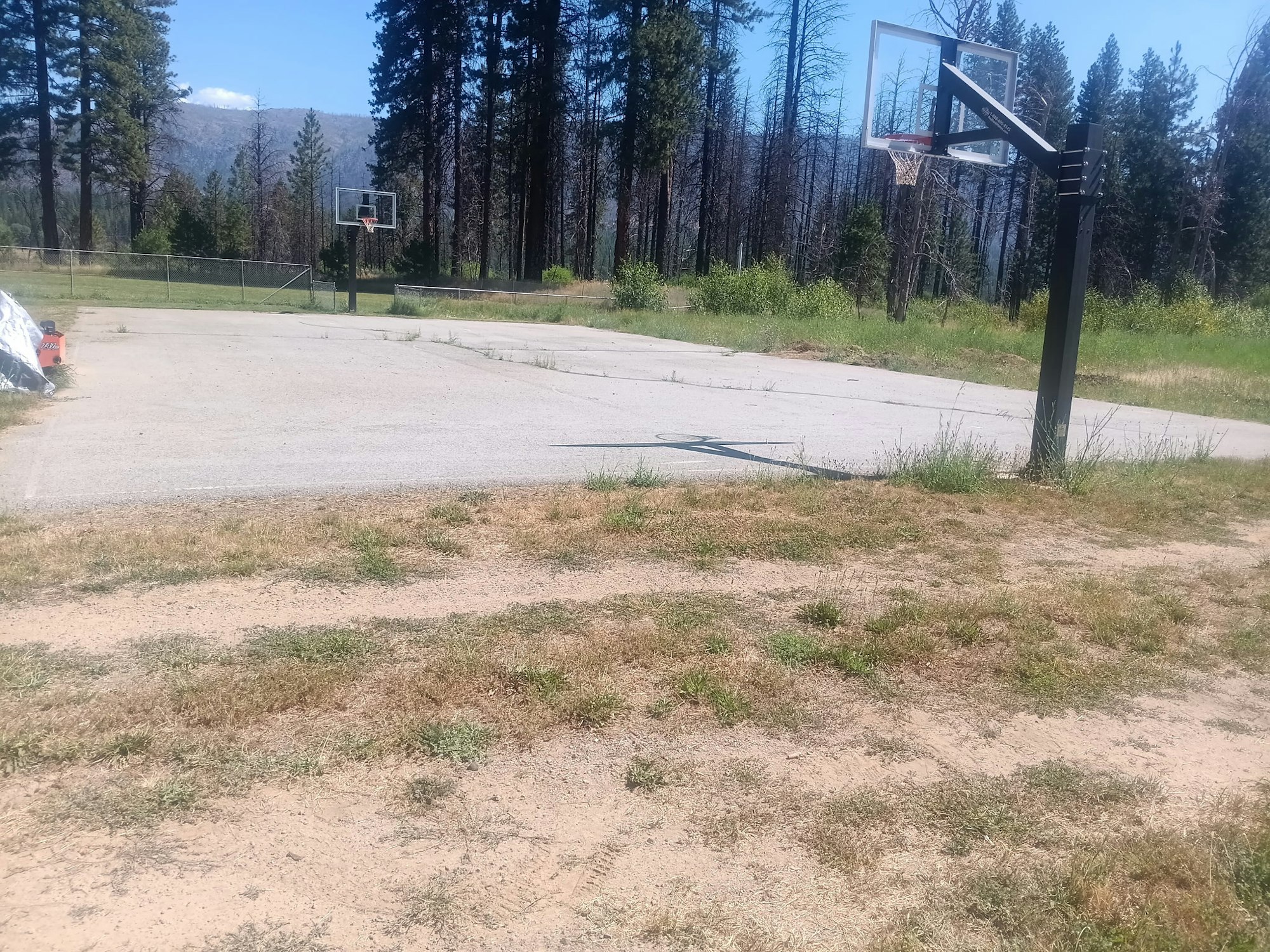 Outdoor basketball court with two hoops, surrounded by grass and trees.