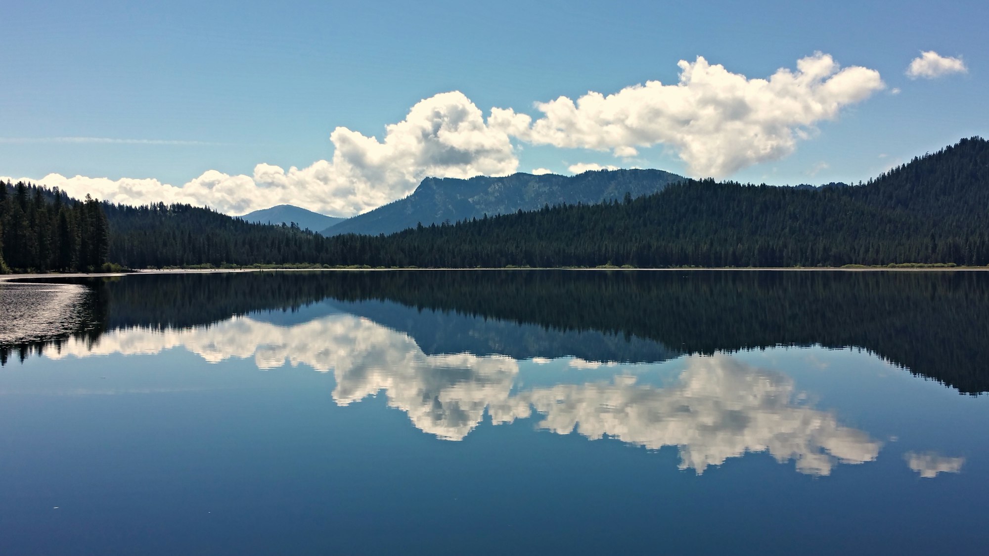 A serene lake reflects mountains and clouds under a blue sky, surrounded by dense forests.