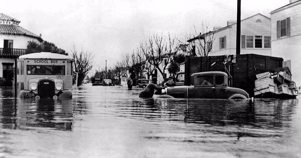A flooded street scene shows a school bus and vehicles partially submerged, with people navigating through the water.