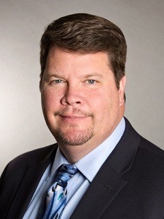 Caucasian man on light gray background wearing dark suit and blue dress shirt and tie.