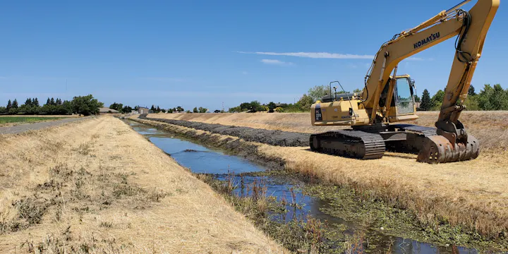 An excavator working alongside a shallow canal in a dry landscape, with sparse vegetation and a clear blue sky.