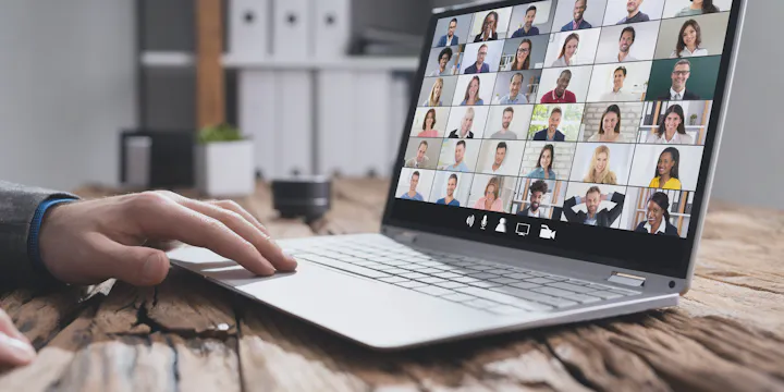 A laptop on a wooden desk displays a video call with multiple participants visible on the screen.