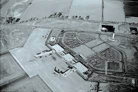 An aerial view of a large industrial or commercial complex surrounded by agricultural land, in black and white.