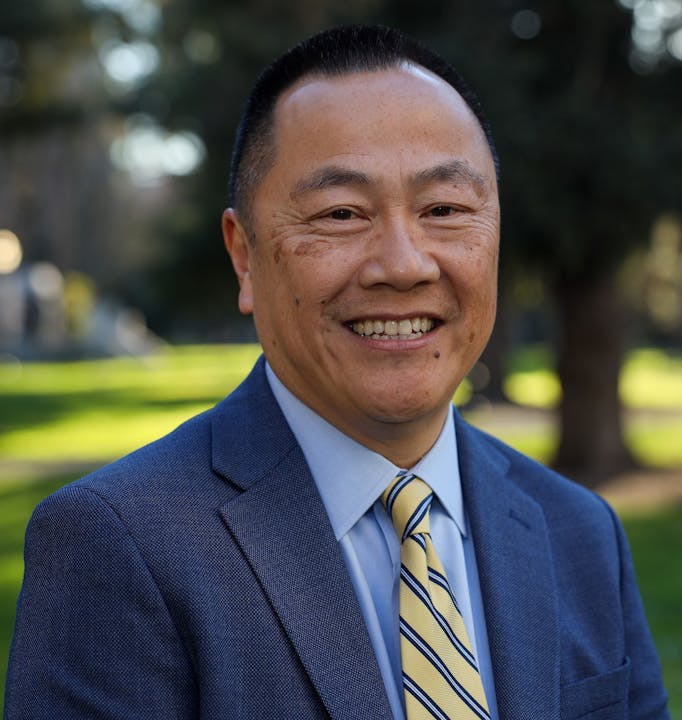 A smiling man in a suit and tie stands outdoors with a blurred green background, exuding confidence and approachability.