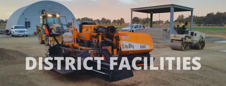 The image shows construction equipment, including a road paver and a roller, in a work area labeled "DISTRICT FACILITIES."