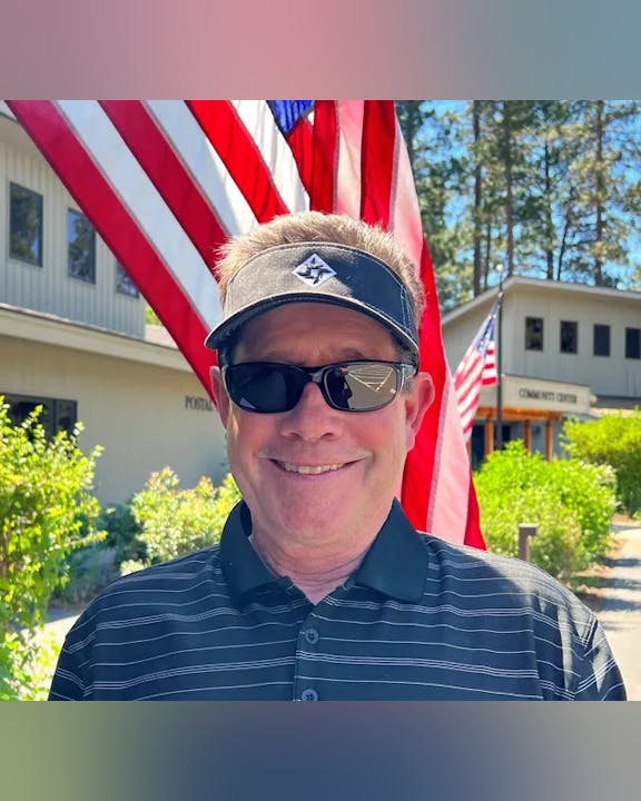A person wearing sunglasses and a visor, smiling in front of American flags and a building labeled "Community Center."