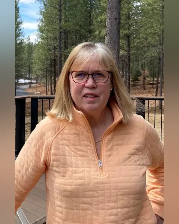 A woman wearing an orange sweater stands outdoors, surrounded by trees, with a road visible in the background.