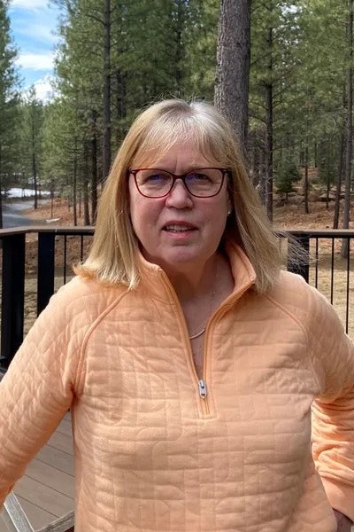 A woman wearing an orange sweater stands outdoors, surrounded by trees, with a road visible in the background.