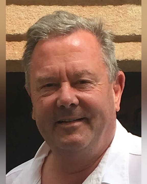 Jay Hamachek, smiling in front of a light brown wall.