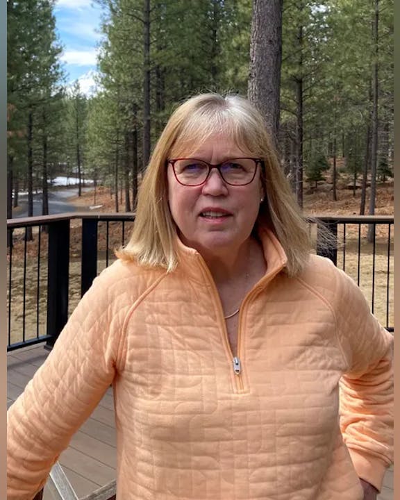 A person wearing glasses and a peach-colored jacket stands on a deck in a forested area.