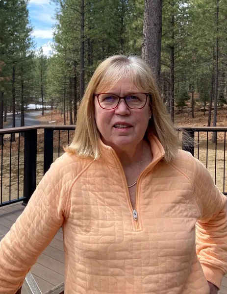 A person wearing glasses and a peach-colored jacket stands on a deck in a forested area.