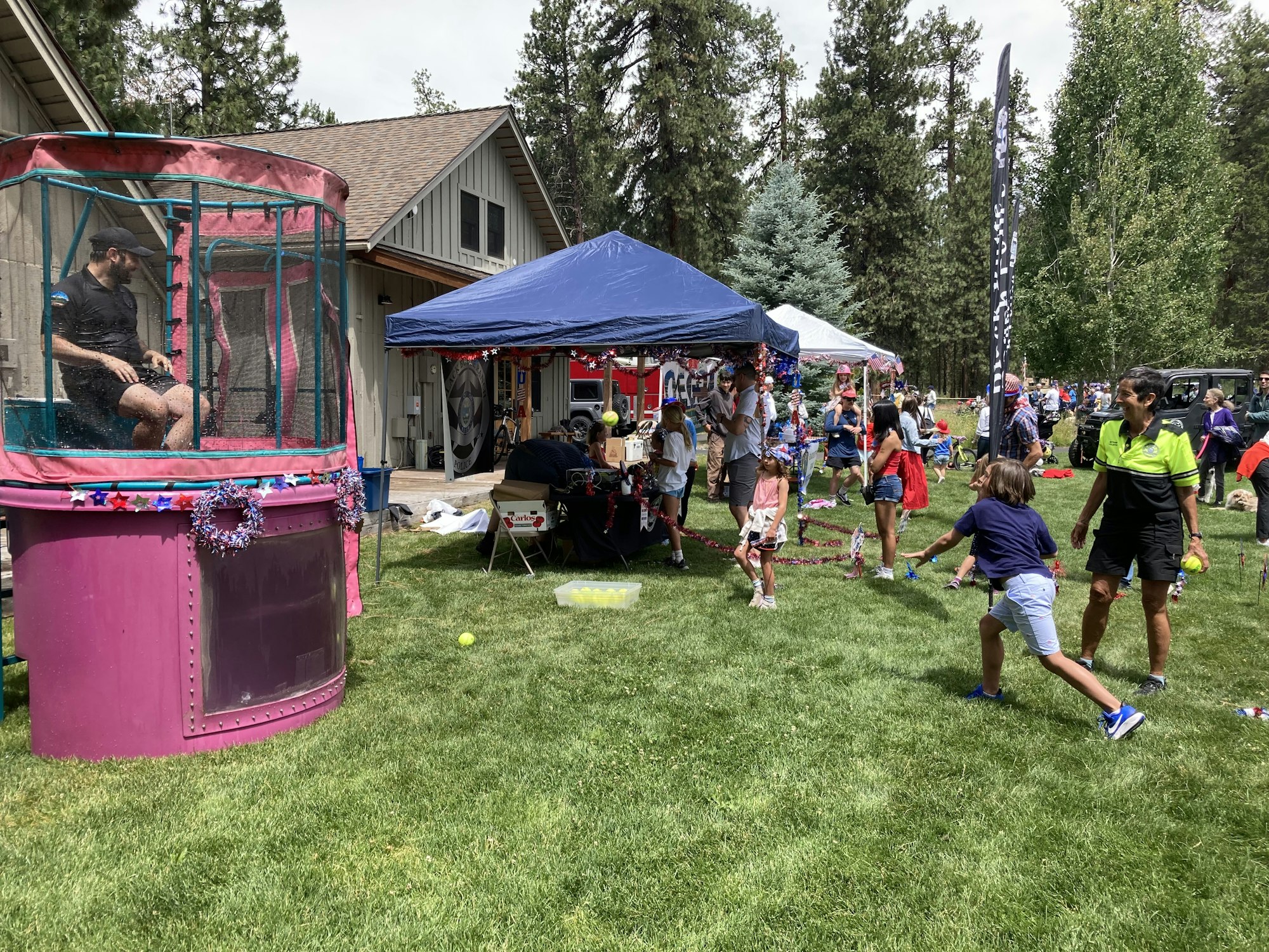A summer event with kids playing, a dunk tank, and festive decorations in a grassy area near a house and tents.