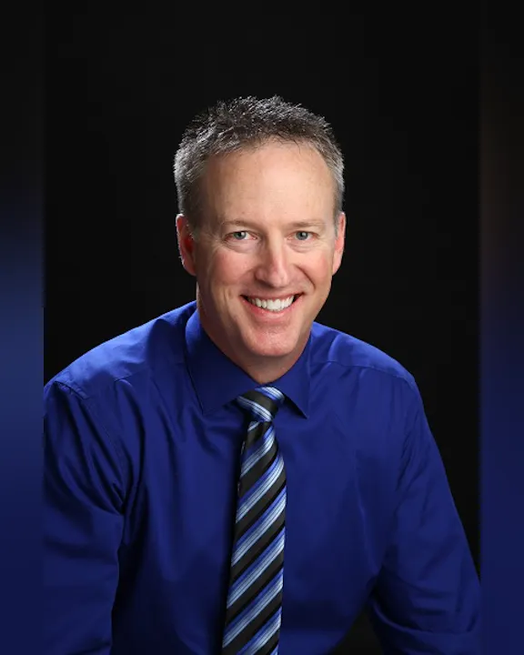 The image features a smiling man in a blue shirt and striped tie, with a black background.