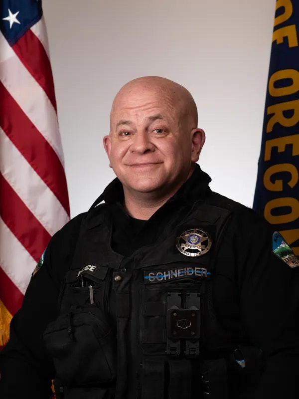 A person in a police uniform with an American flag and an Oregon state flag in the background.