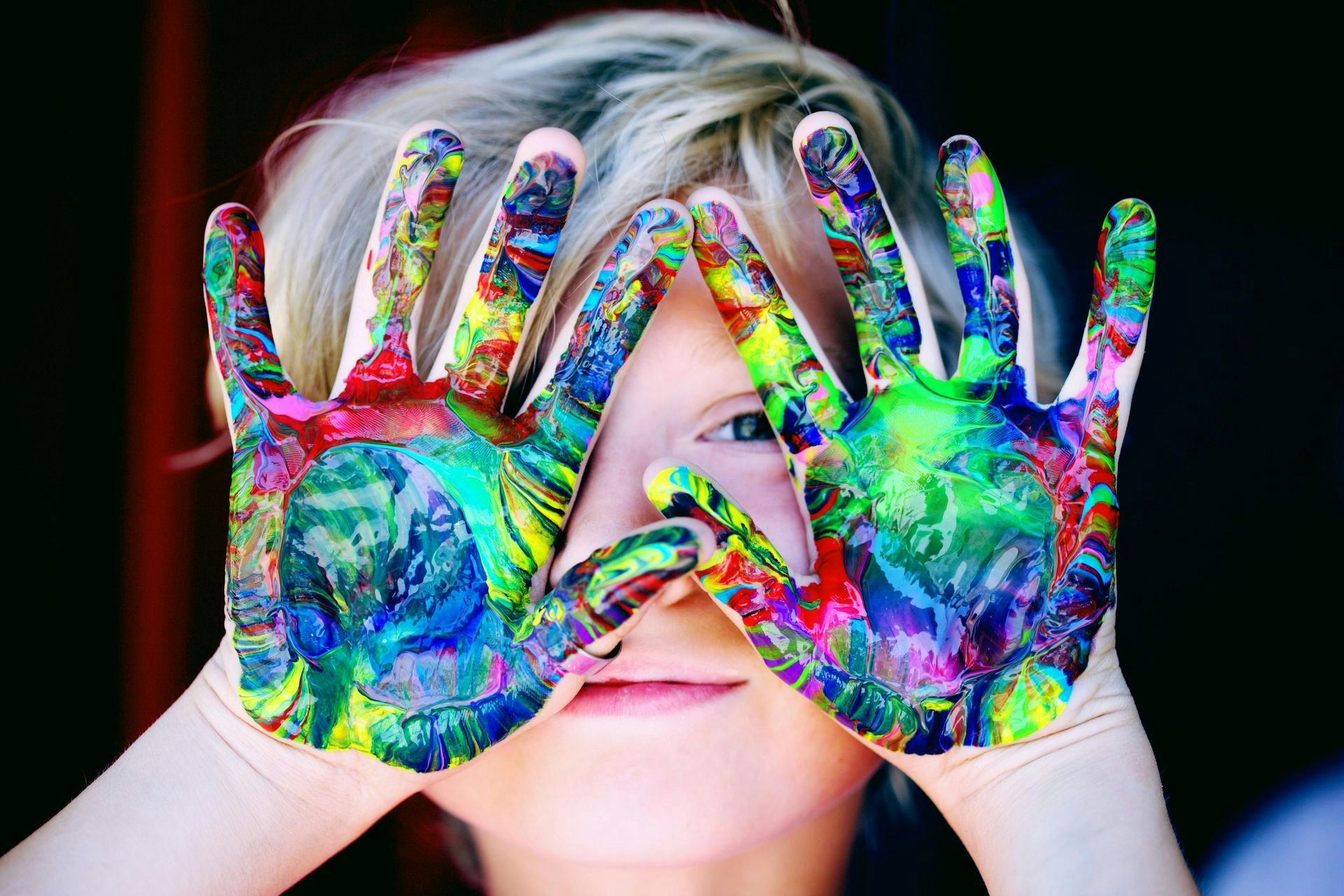 Child with colorful painted hands covering face, peeking through fingers.
