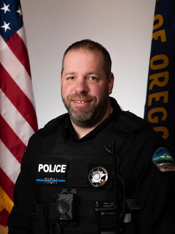 A police officer in uniform, with a U.S. flag and an Oregon flag in the background.