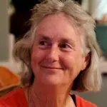 The image shows a smiling woman with gray hair, wearing an orange top, in a warm and inviting indoor setting.