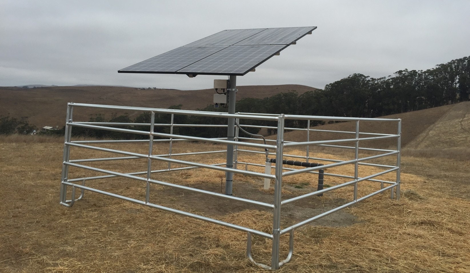 A solar panel mounted on a pole, enclosed by metal fencing, likely for agricultural or water management purposes in a rural area.