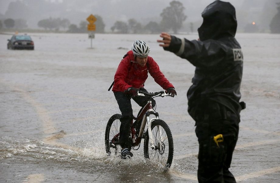 A cyclist in a red jacket navigates through floodwaters, while a police officer gestures for caution in heavy rain.