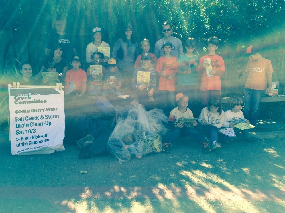 A community group, including many children, gathers for a Fall Creek & Storm Drain clean-up event, promoting environmental action.