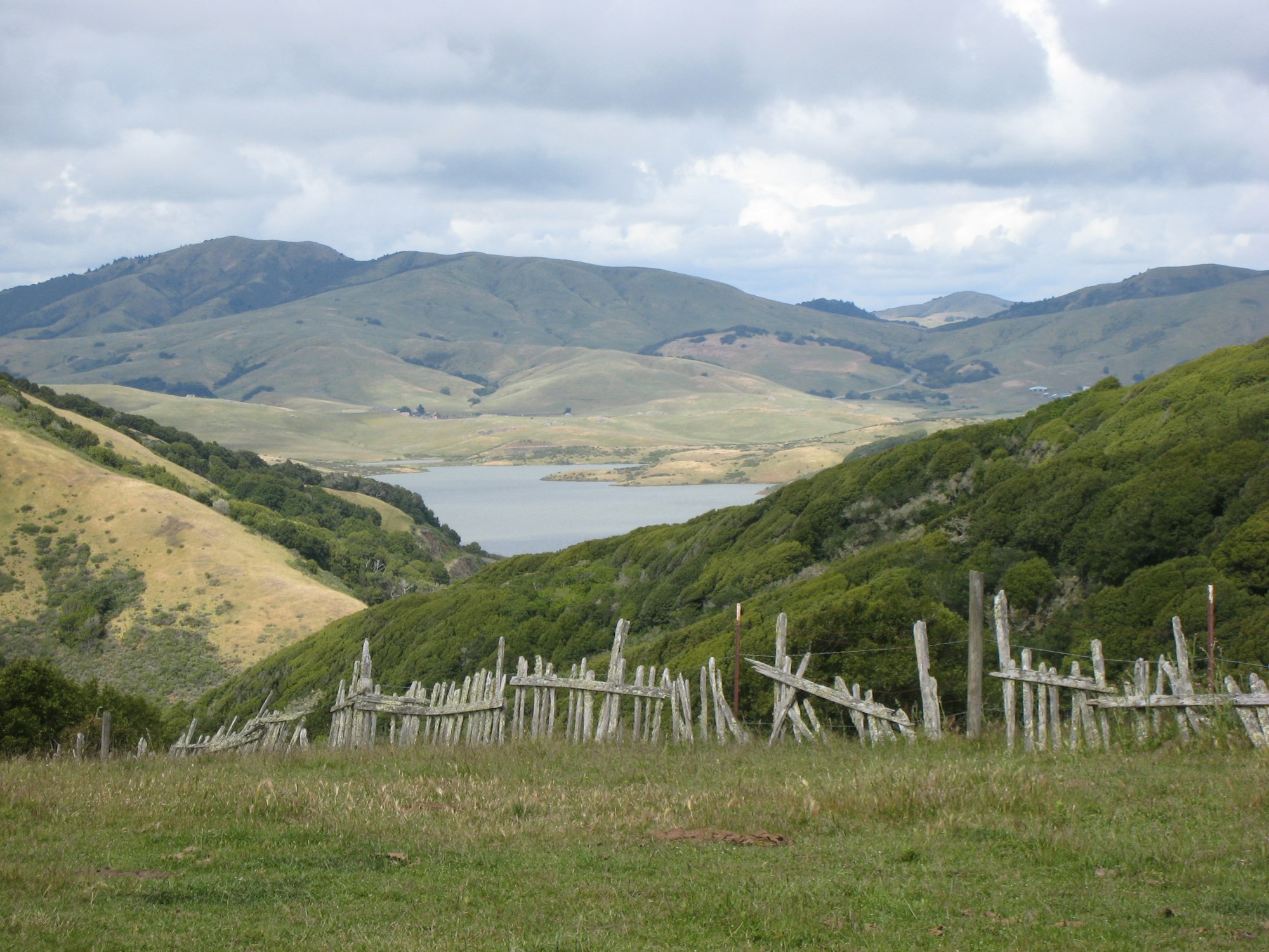 A scenic landscape featuring rolling hills, a serene lake, and a rustic wooden fence under a cloudy sky.