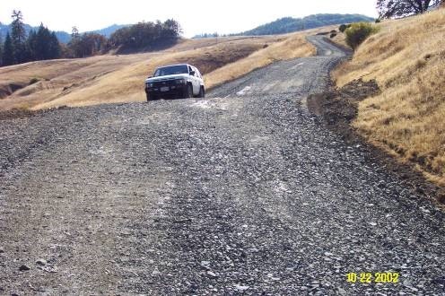 A gravel road winding through a hilly landscape, with a vehicle parked along the side. Sunlit scenery in the background.