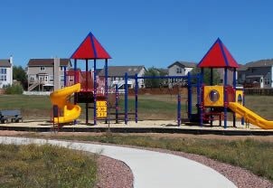 A colorful children's playground with slides and climbing structures, set against a clear sky and suburban backdrop.