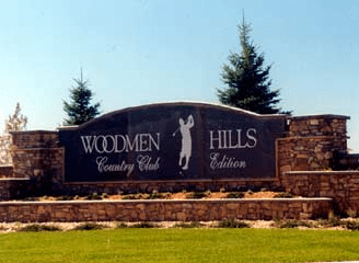Sign for "Woodmen Hills Country Club Edition" with a golfer silhouette, set on a stone wall, with trees and grass.