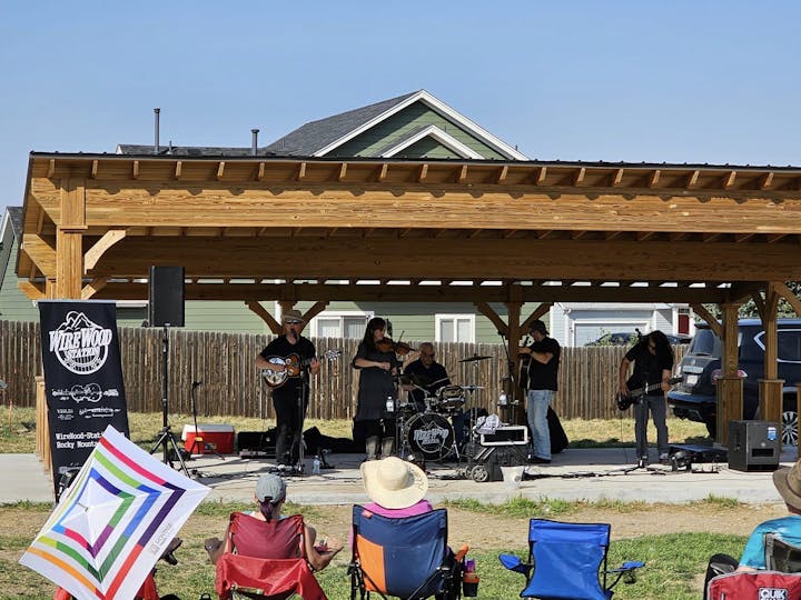 Outdoor live music event with a band performing on a stage and audience seated in portable chairs.