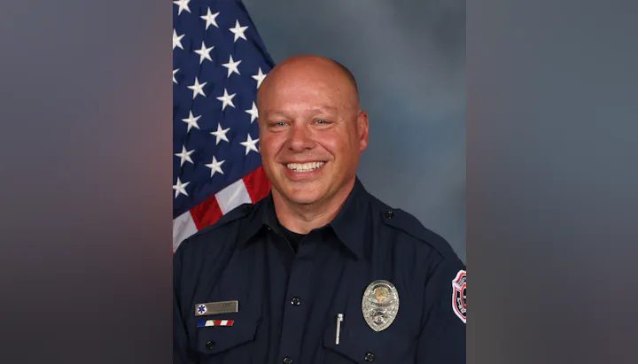 The image shows a smiling firefighter in uniform, with an American flag in the background. He has a badge and name tag visible.