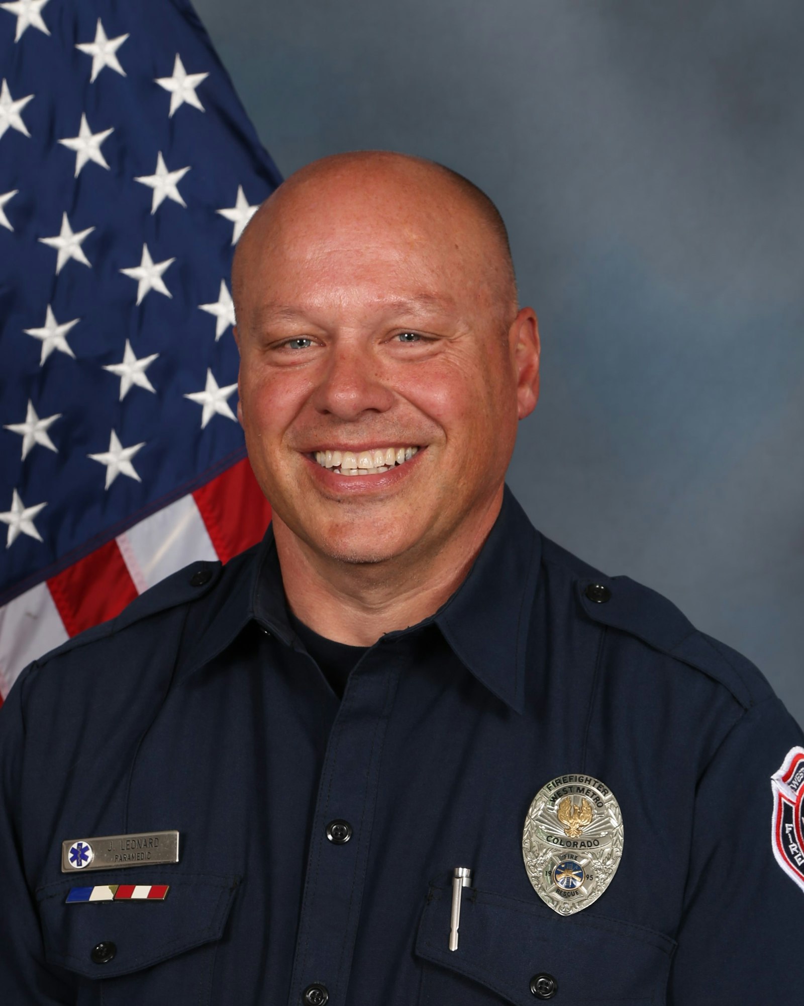 The image shows a smiling firefighter in uniform, with an American flag in the background. He has a badge and name tag visible.