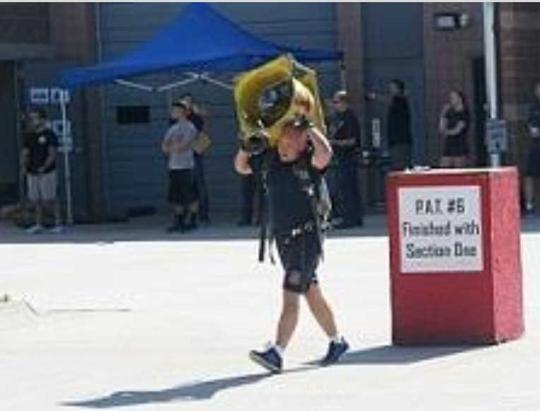 A person carrying a heavy yellow object outdoors near a red sign that reads "RAT #6 Finished with Section One." Several people are in the background.