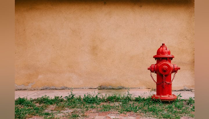 A red fire hydrant against a yellow wall with grass below it.