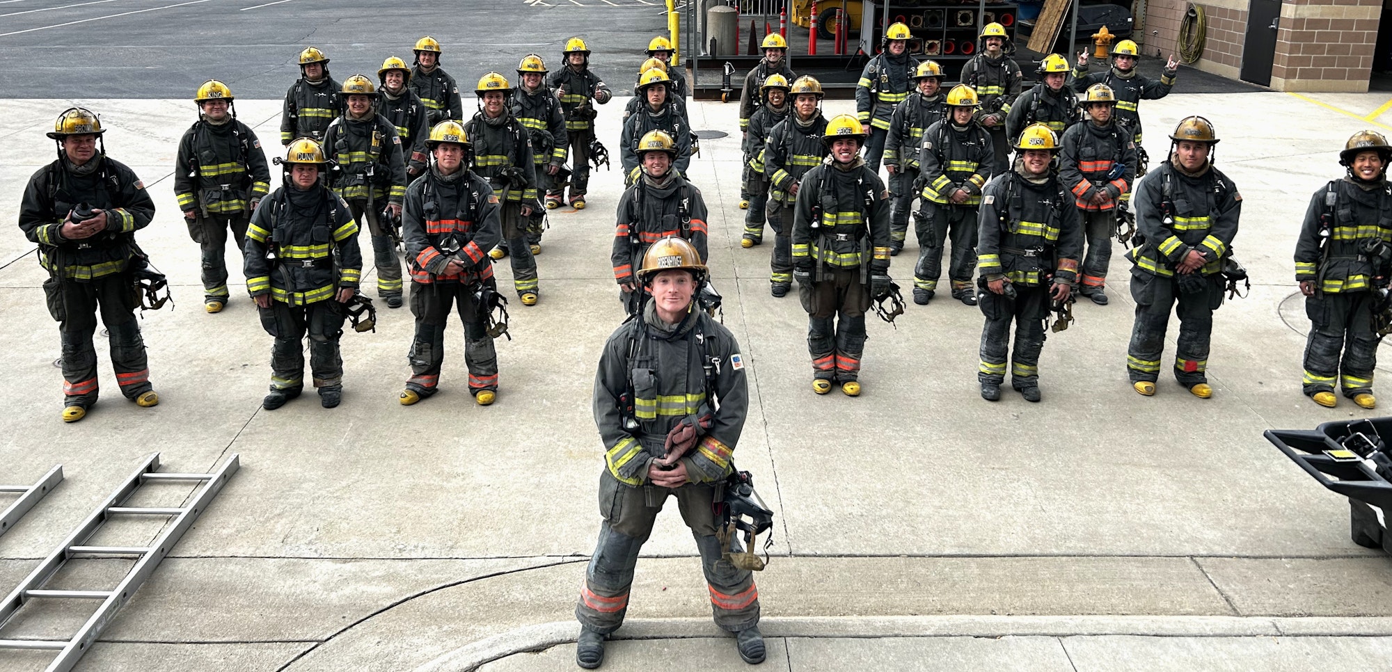 A group of firefighters in uniform and helmets stand together on a concrete surface, facing the camera.