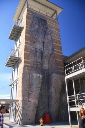A tall rock climbing wall on a building with metal stairs and people nearby.