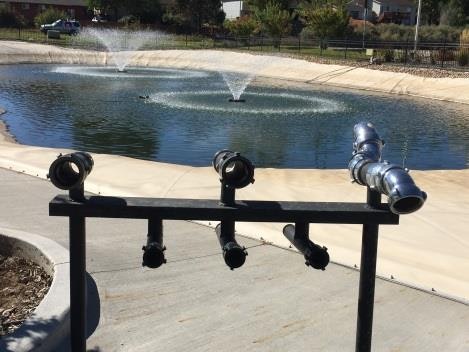 A pond with two fountains and a foreground featuring several metallic pipes on a stand.