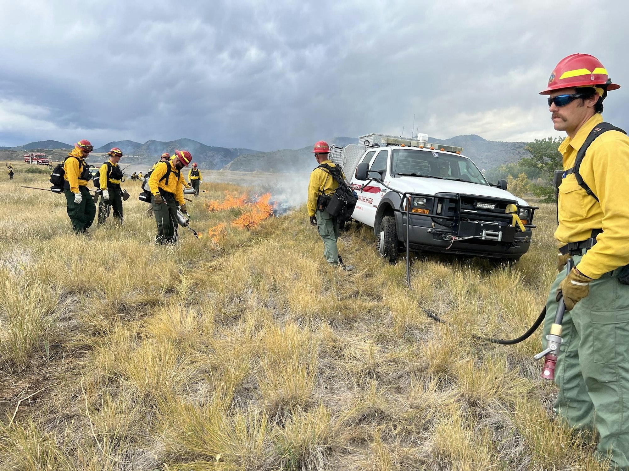 West Metro firefighters working prescribed burn at Bear Creek Lake Park. Picture shows a line of firefighters using drip torches to light the grass, and one firefighter standing by with a charged hose line.