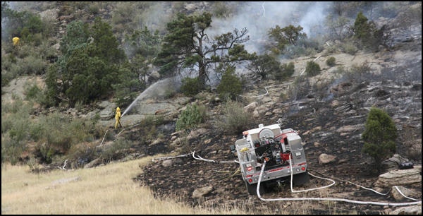 Wildland fire with brush truck surrounded by burned grass and firefighters in the background, using a hose to extinguish flames.